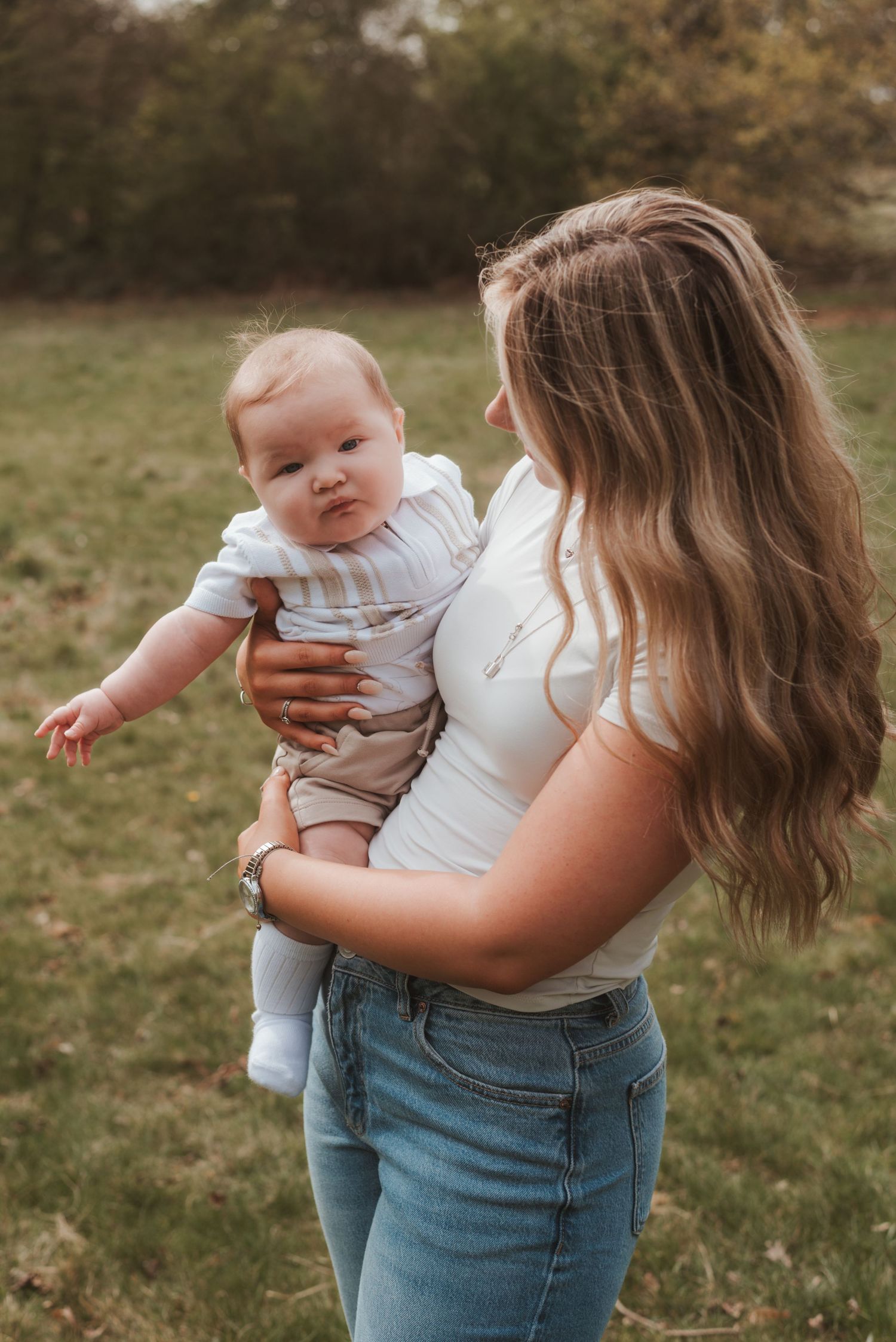 Baby in white outfit reaching out while being held outdoors in autumn setting.