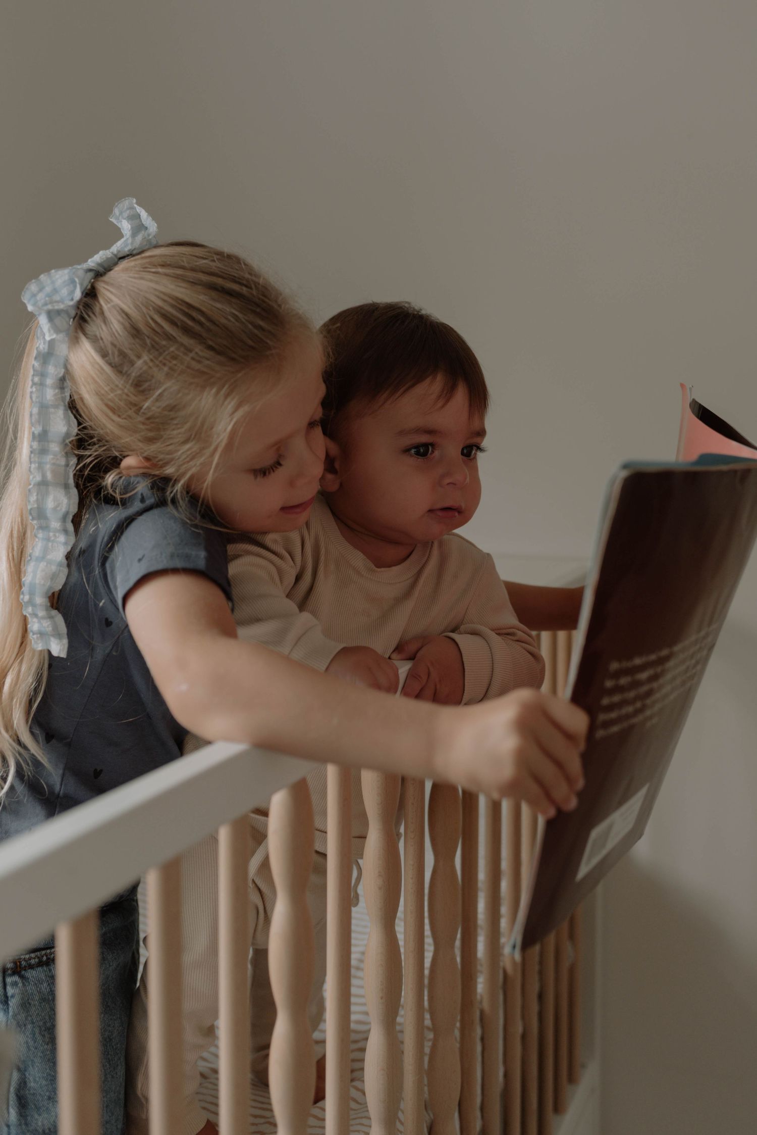 Two children peek over the railing of a wooden crib together in a dimly lit room.