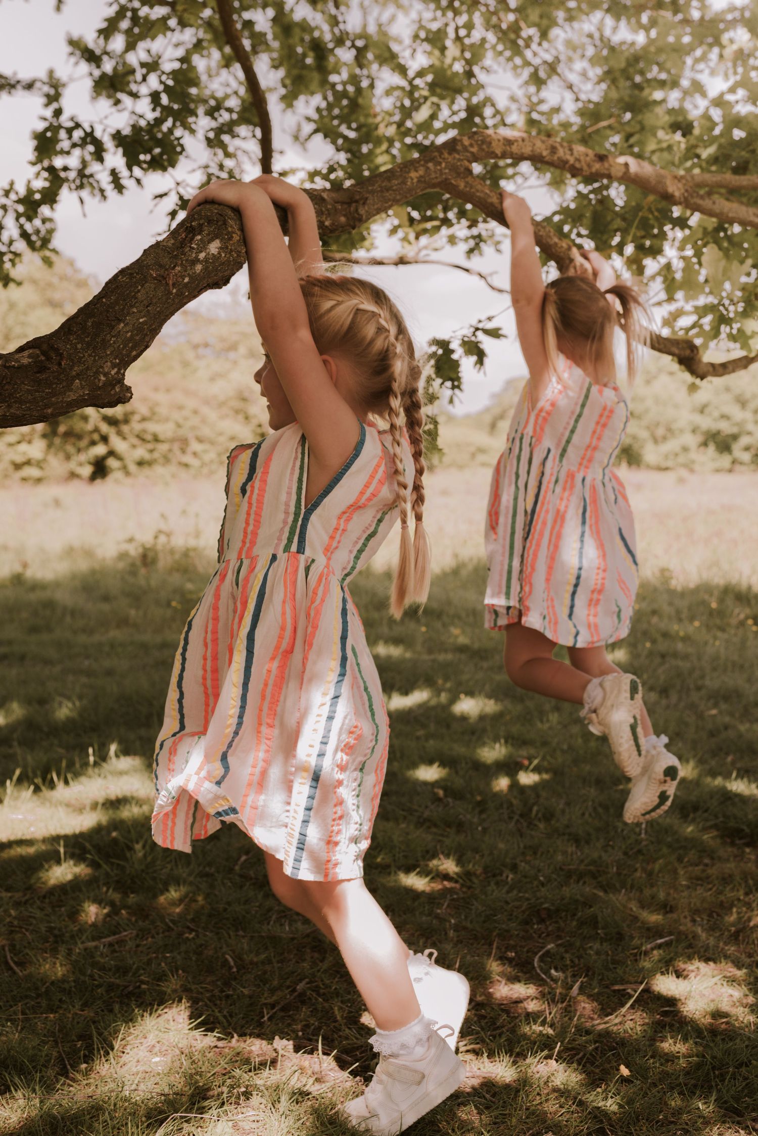 Two children in matching striped dresses swing from a tree branch on a sunny day.