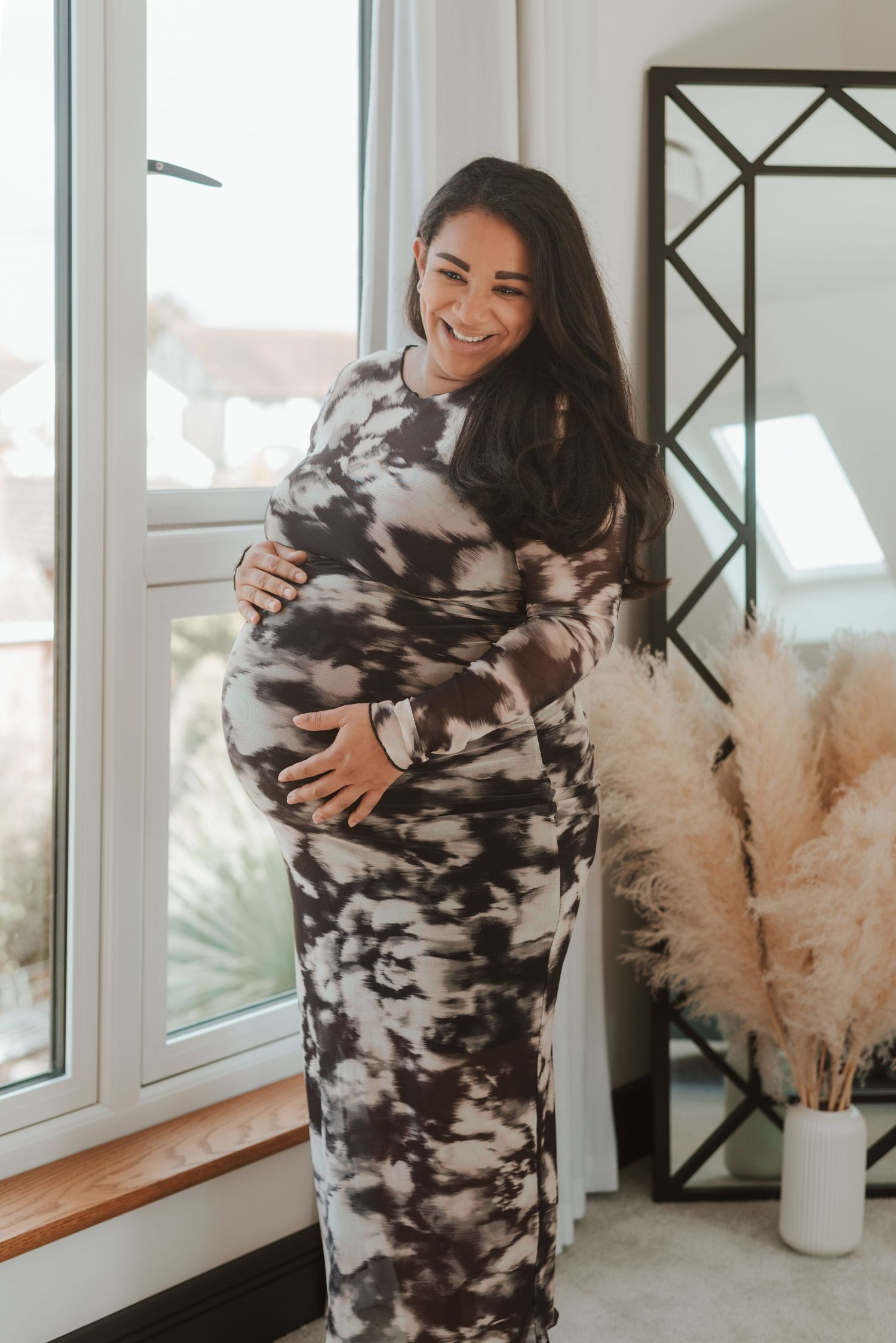 A maternity photo featuring a black and white camo print dress near bright windows and pampas grass decor.