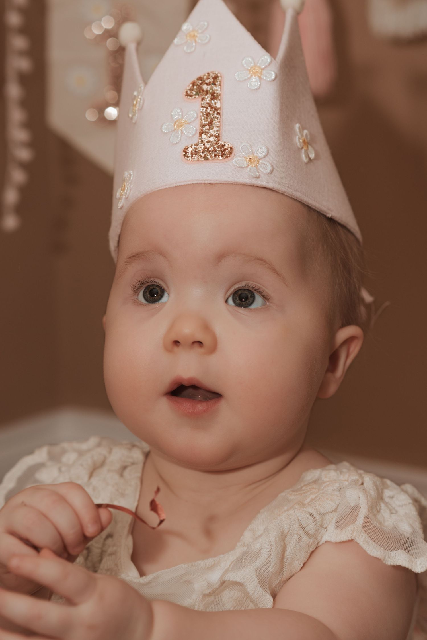 Baby wearing a decorative first birthday crown and white lace outfit smiles with bright eyes.