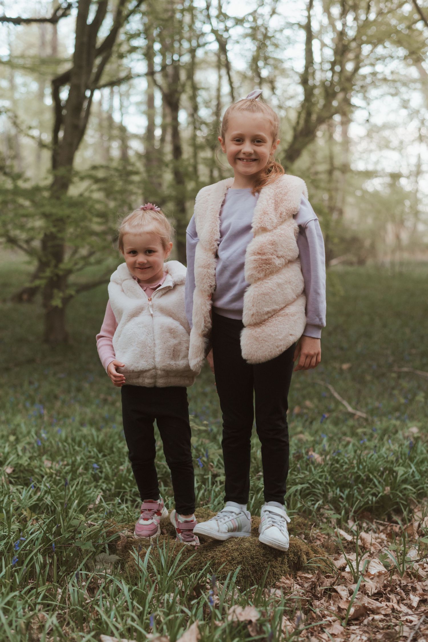 Two children in matching fur vests standing together in a wooded setting.