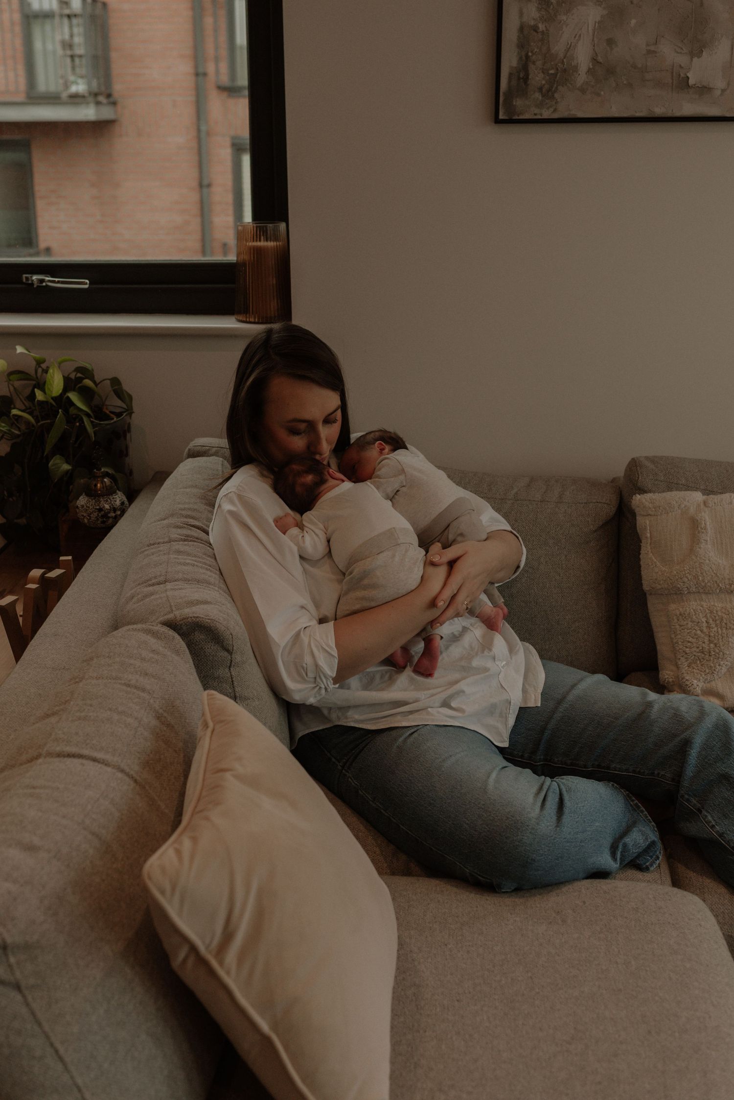A tender moment on a beige couch as someone cradles a sleeping baby in a cozy living room with natural lighting.