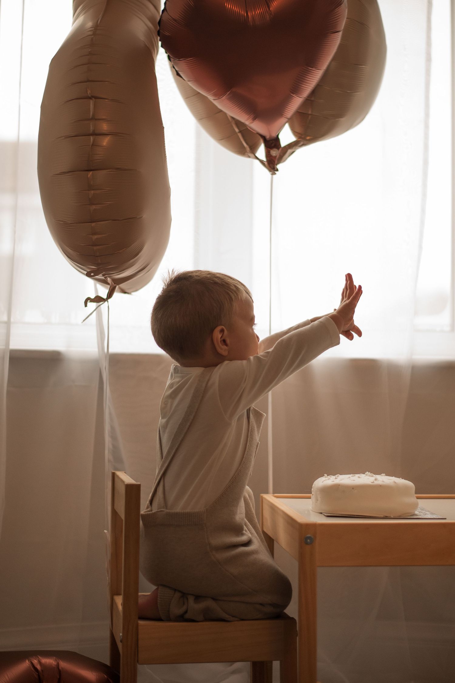 Baby in white reaching for brown balloons by window in sunlit room.