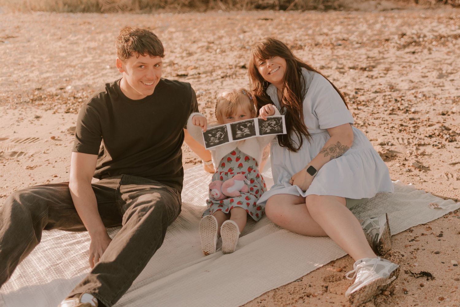 A family maternity photo session on a beach blanket with ultrasound photos displayed.