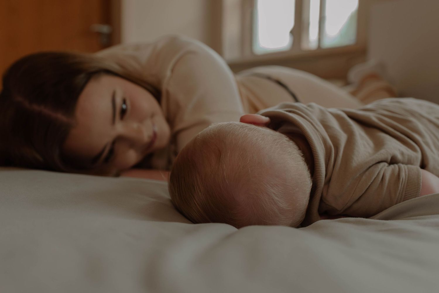 A parent and baby lying together on white bedding in warm natural light from a nearby window.