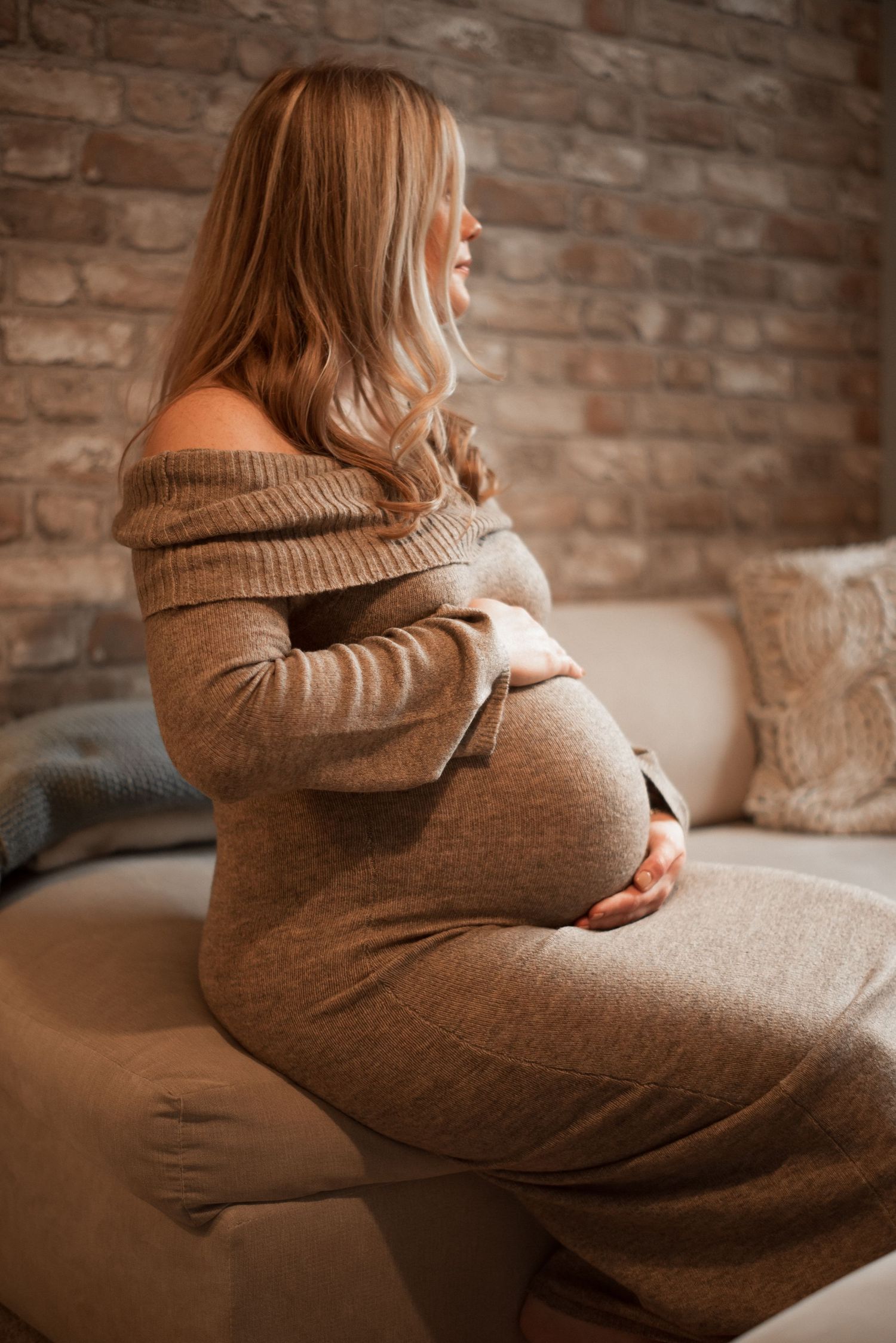 Pregnant woman in beige sweater dress sitting sideways on couch.
