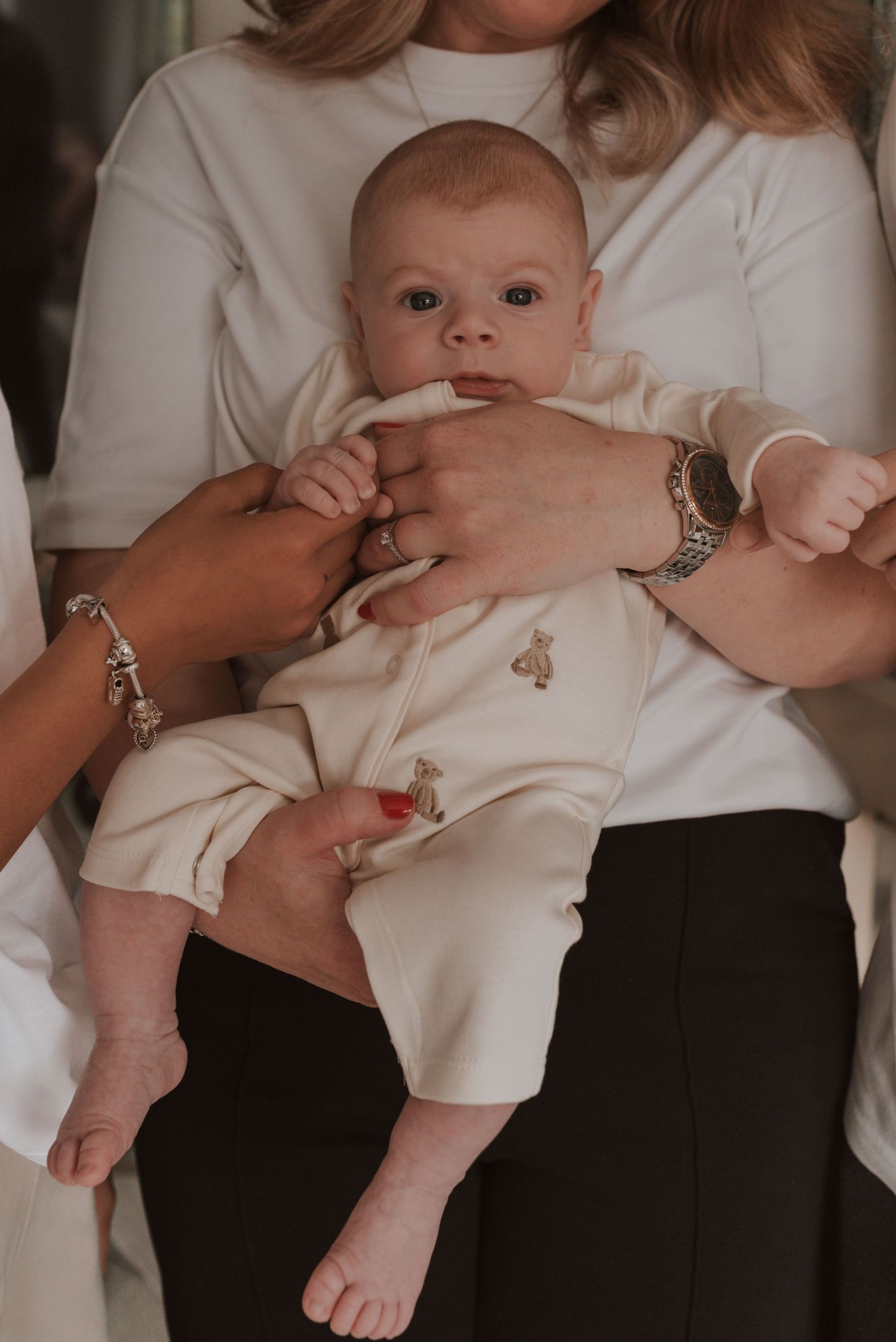 An infant wearing a white onesie with tiny bear prints is held by someone wearing a white shirt and silver bracelets.