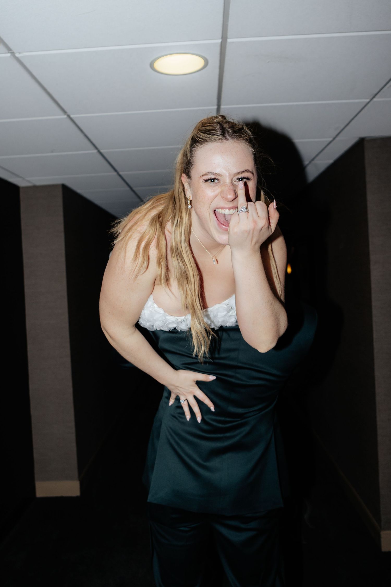 Excited blonde woman in green satin dress showing ring, laughing joyfully in a dimly lit hallway.