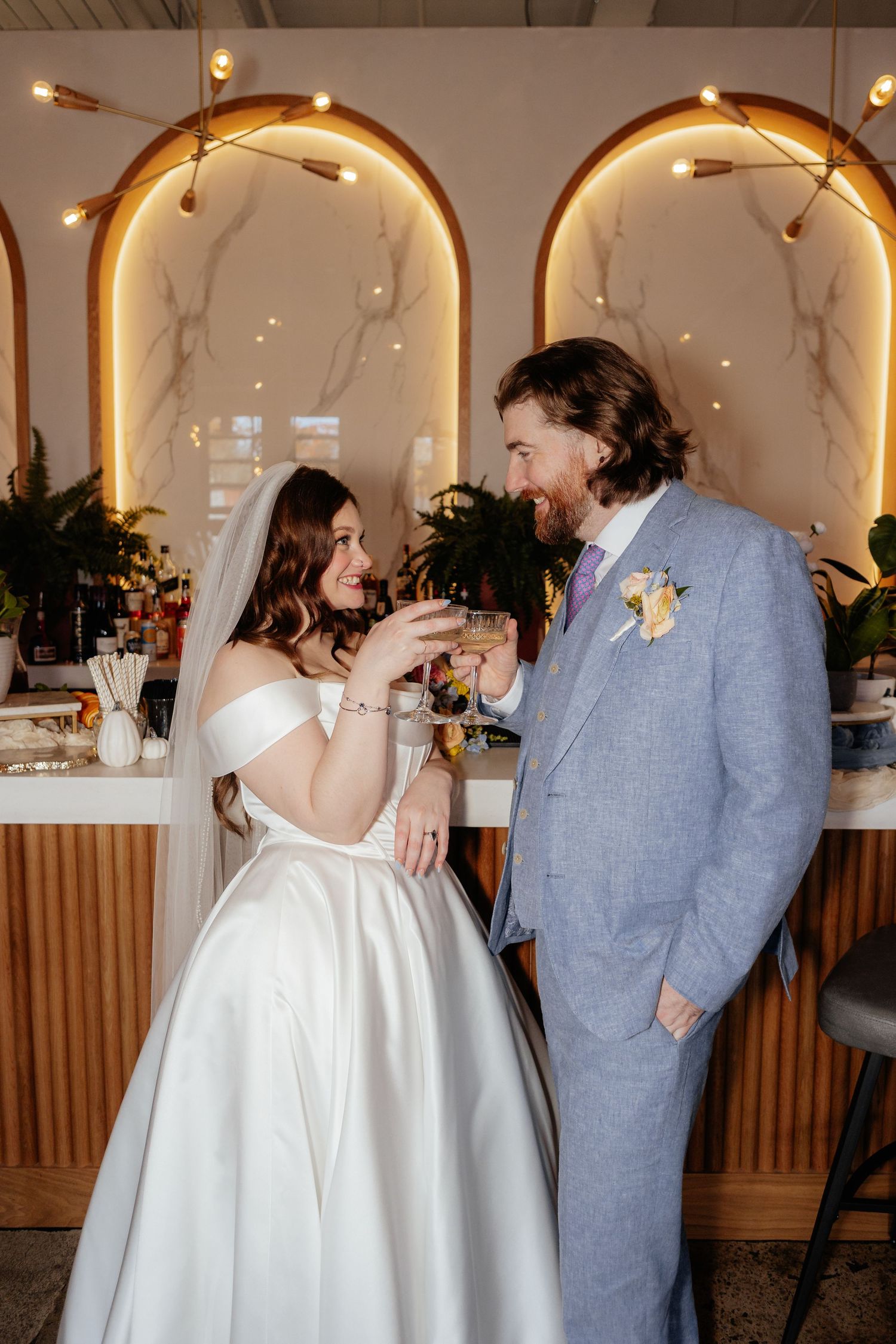 Bride and groom toasting with champagne glasses at elegant wedding venue with arched marble bar backdrop.