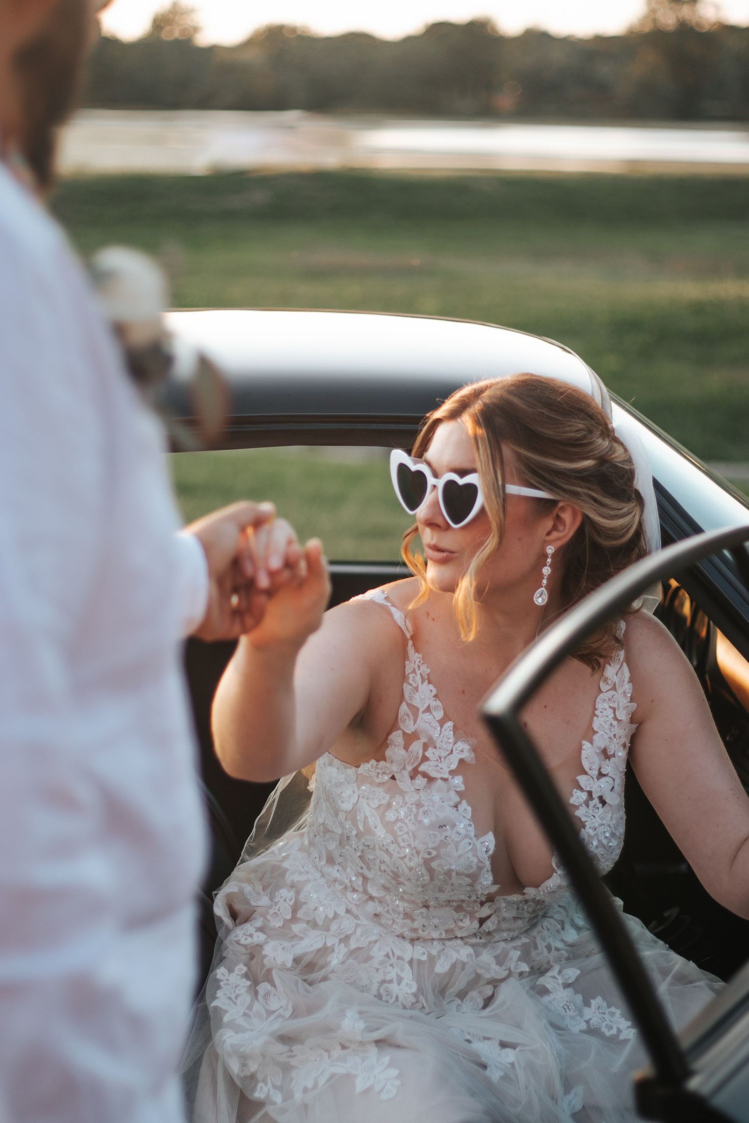 Bride in lace dress and sunglasses sitting in convertible car during sunset.