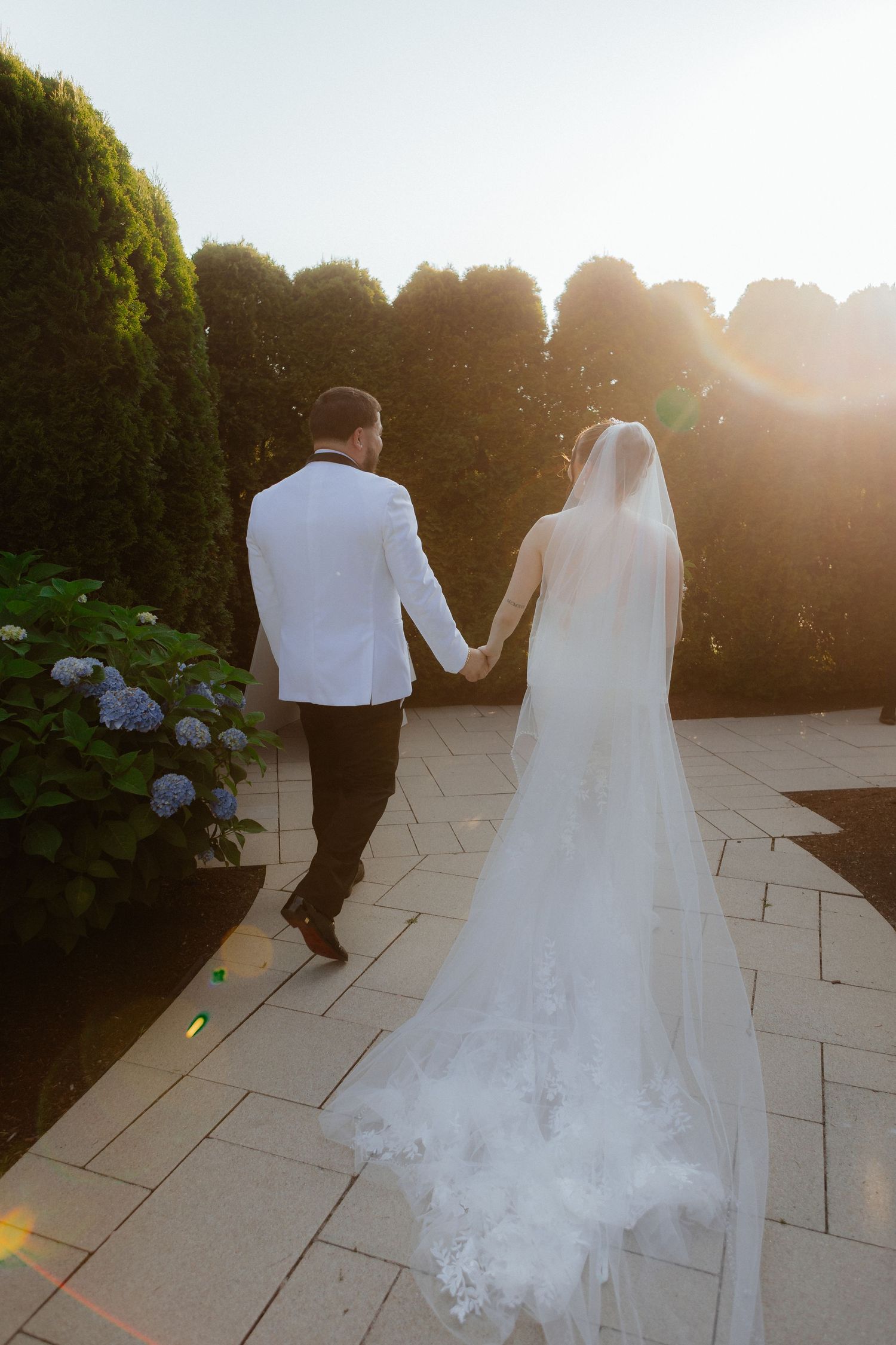 Bride and groom walking hand in hand through a sunlit garden path, bride wearing a lace-trimmed cathedral veil.