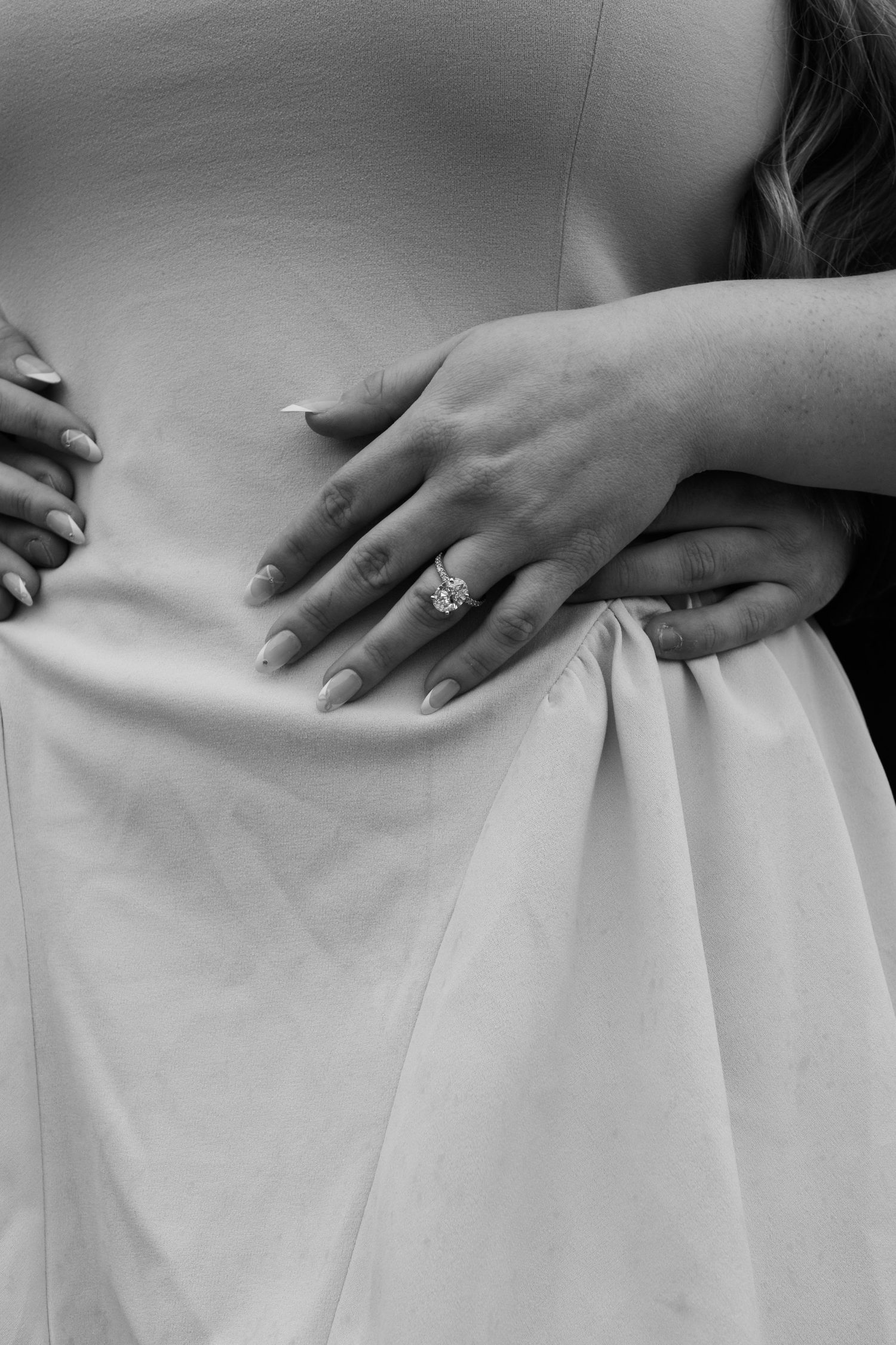 Black and white detail shot of hands and engagement ring against a wedding dress.