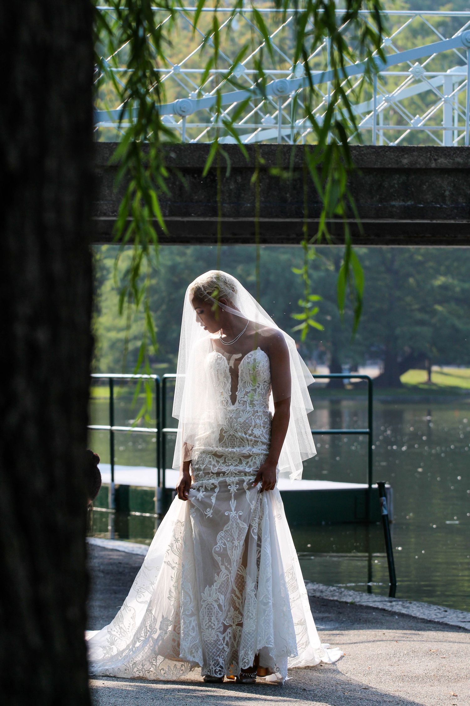 Elegant lace wedding dress silhouette against a waterfront backdrop with hanging willow branches.