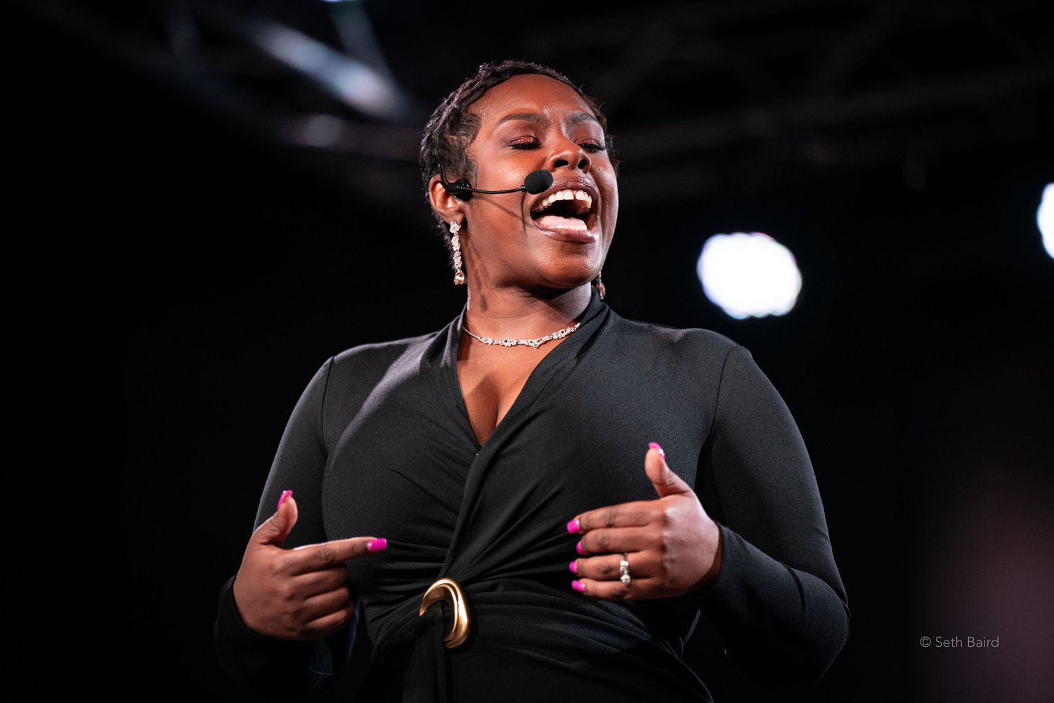 Woman in black wrap dress sings passionately on stage with hands on chest.