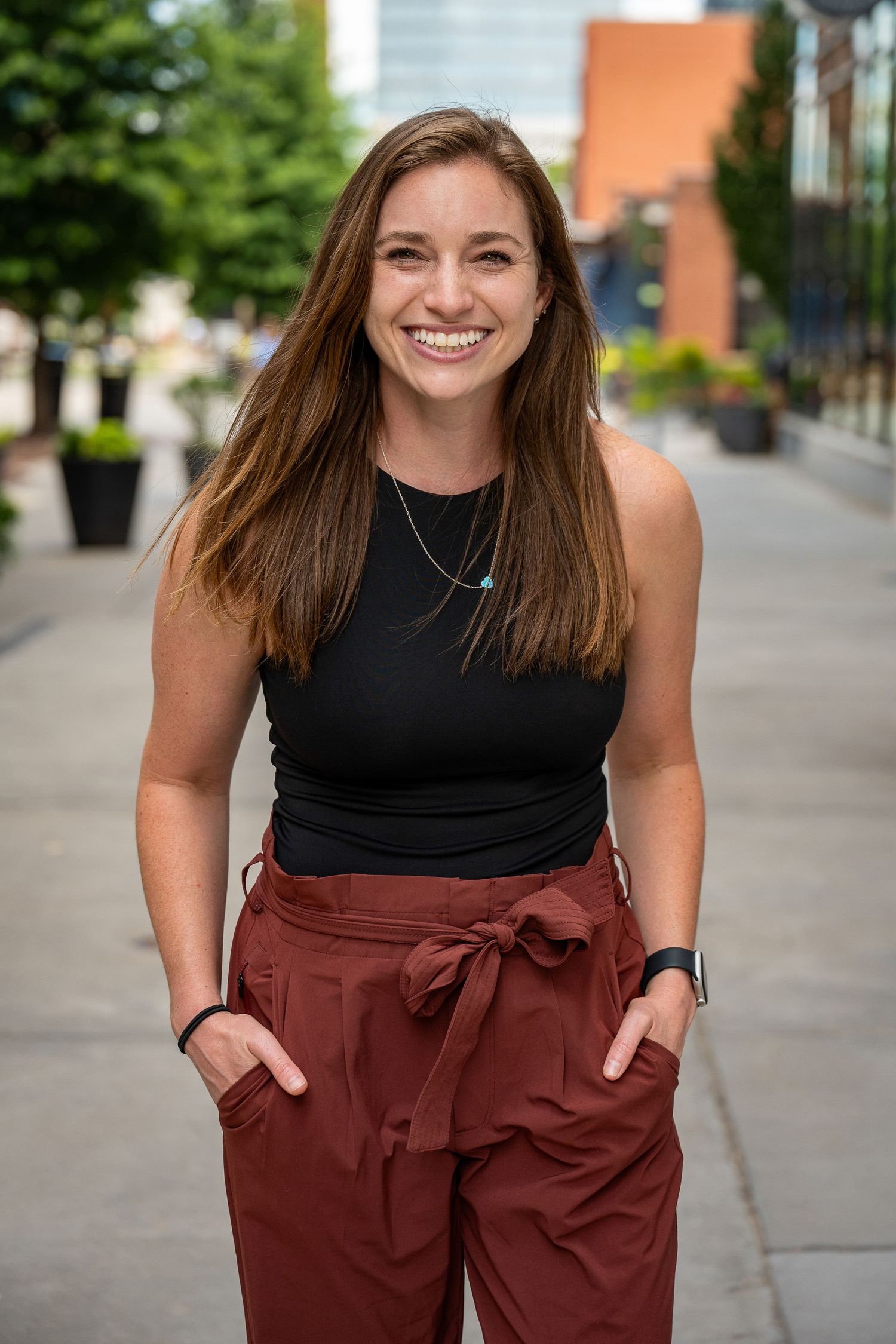 Smiling young woman in black tank top and burgundy tie-waist pants posing outdoors in urban setting.