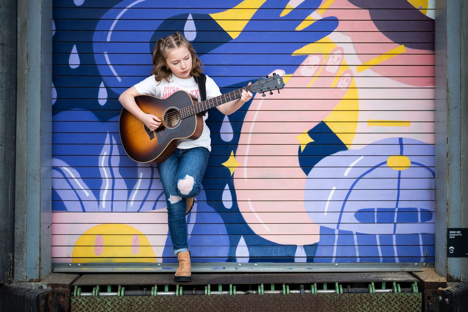 Young girl playing an acoustic guitar in front of a colorful blue and pink mural on a metal door.