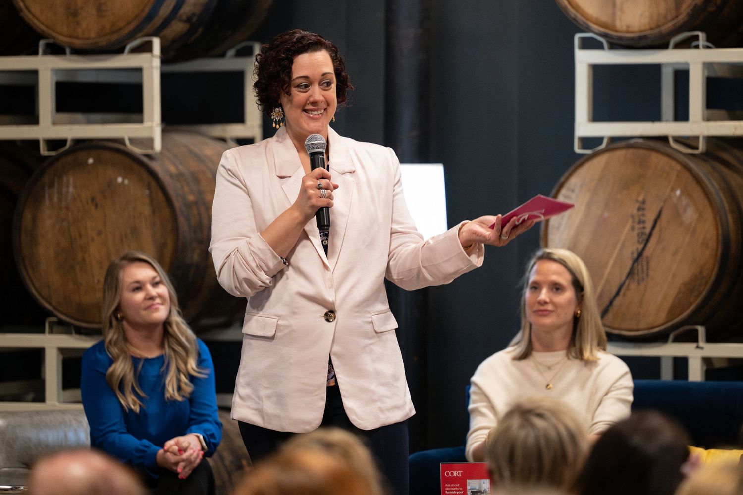 Woman in pink blazer speaks into microphone before seated audience near wooden barrels backdrop.