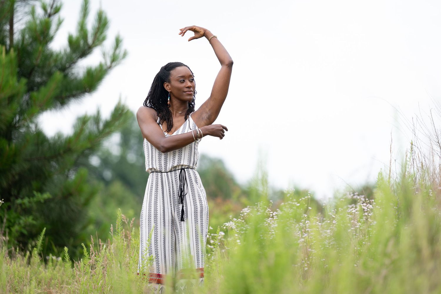 A woman in a striped jumpsuit dances gracefully in a lush green meadow surrounded by wildflowers and trees.