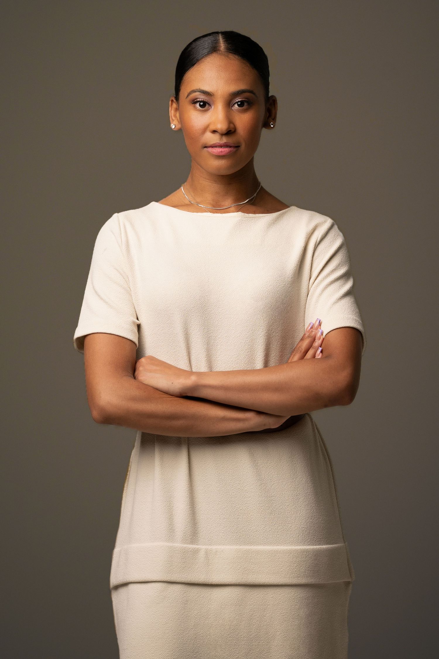 Professional Black woman in cream outfit stands with arms crossed against gray background, exuding confidence.