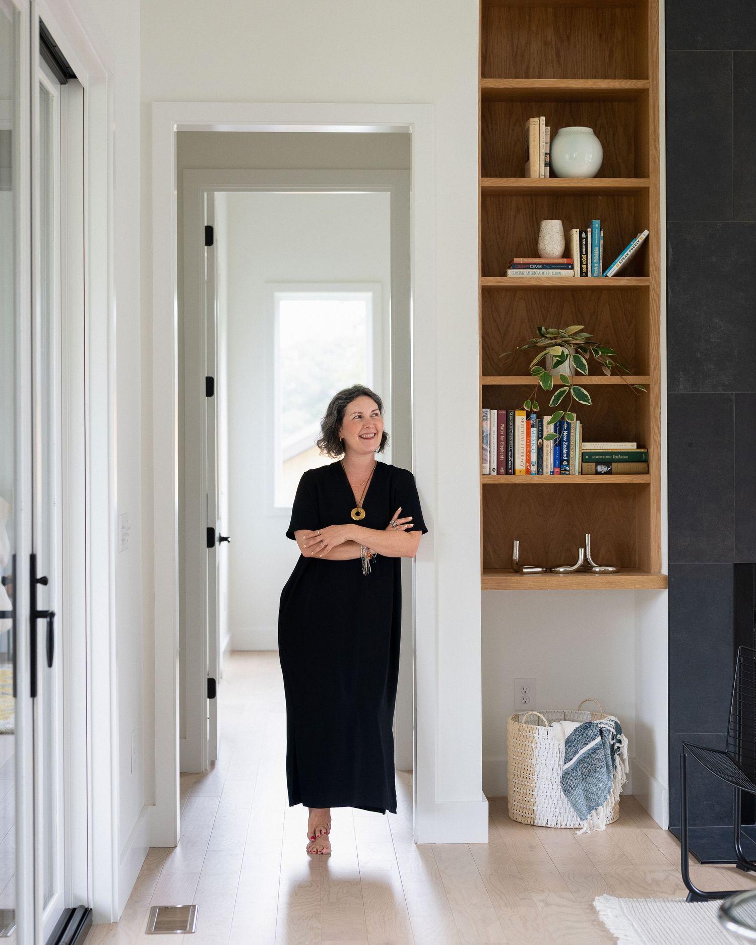 Woman in black maxi dress laughing in modern home interior with wooden built-in bookshelf.