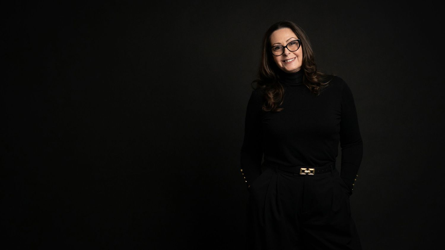 Woman in black turtleneck and glasses poses confidently against dark background in professional portrait photo.
