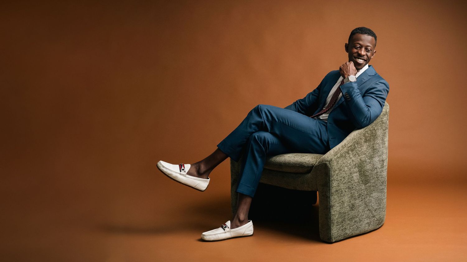Stylish man in blue suit sitting relaxed in chair against warm brown studio background.