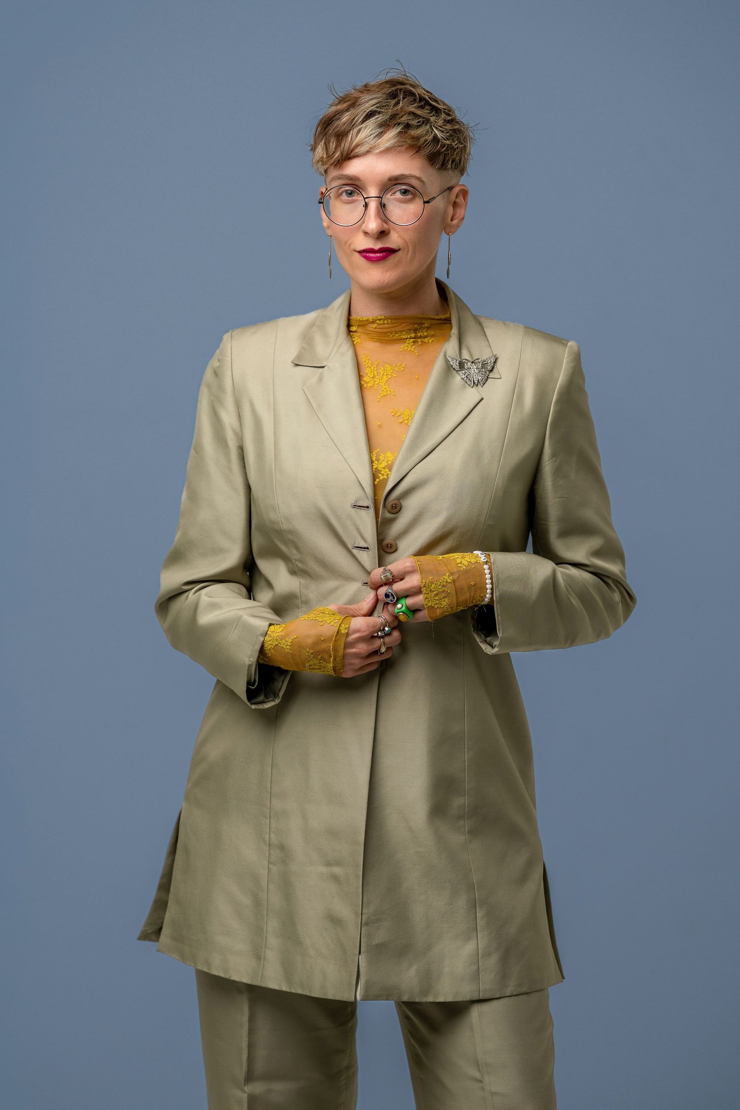 Woman in sage green pantsuit and yellow top with glasses poses confidently against a blue background.