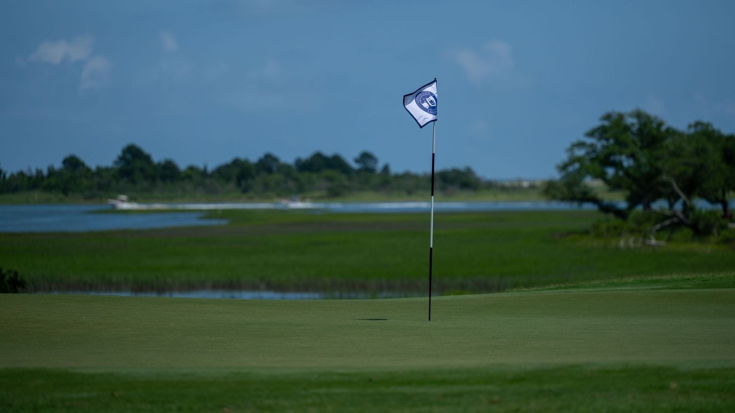 Golf course flag waves near scenic water hazard under cloudy blue sky.