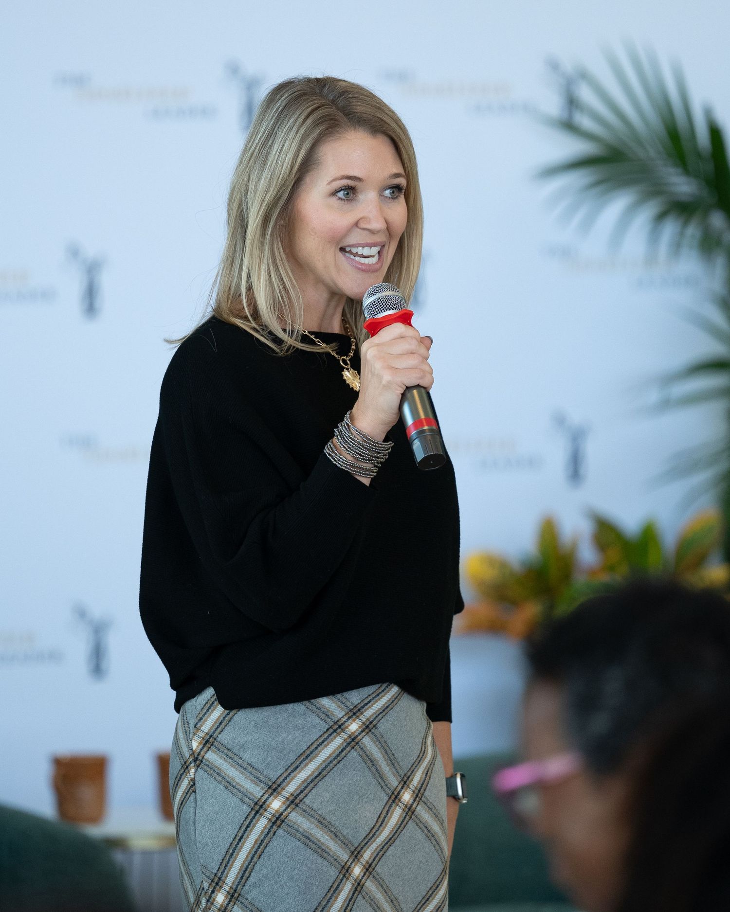 Blonde woman in black sweater and plaid skirt speaking into microphone at a professional event.