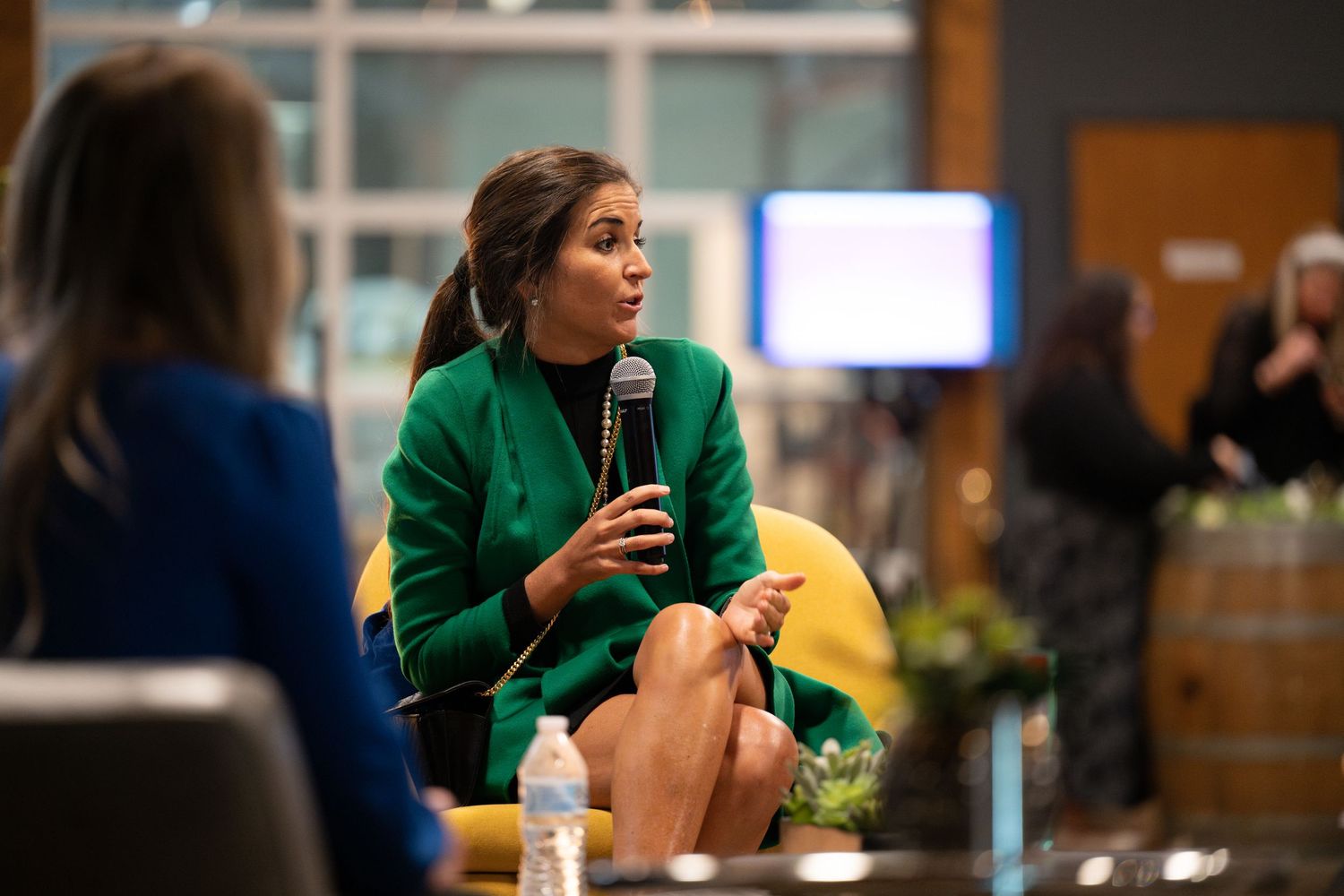Woman in green blazer speaks into microphone while seated at a panel discussion event.