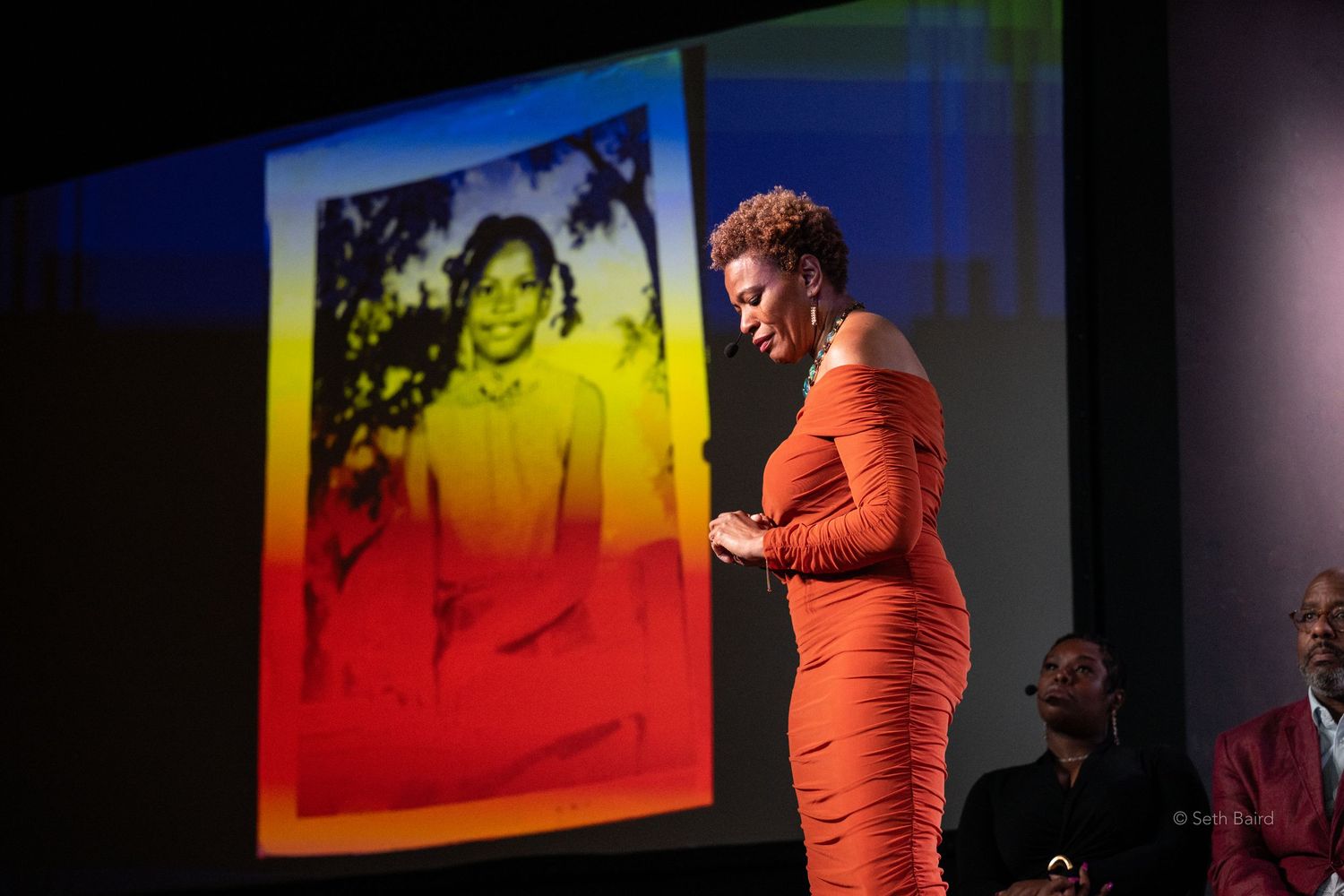 Woman in orange dress presents on stage beside colorful projected portrait photograph.