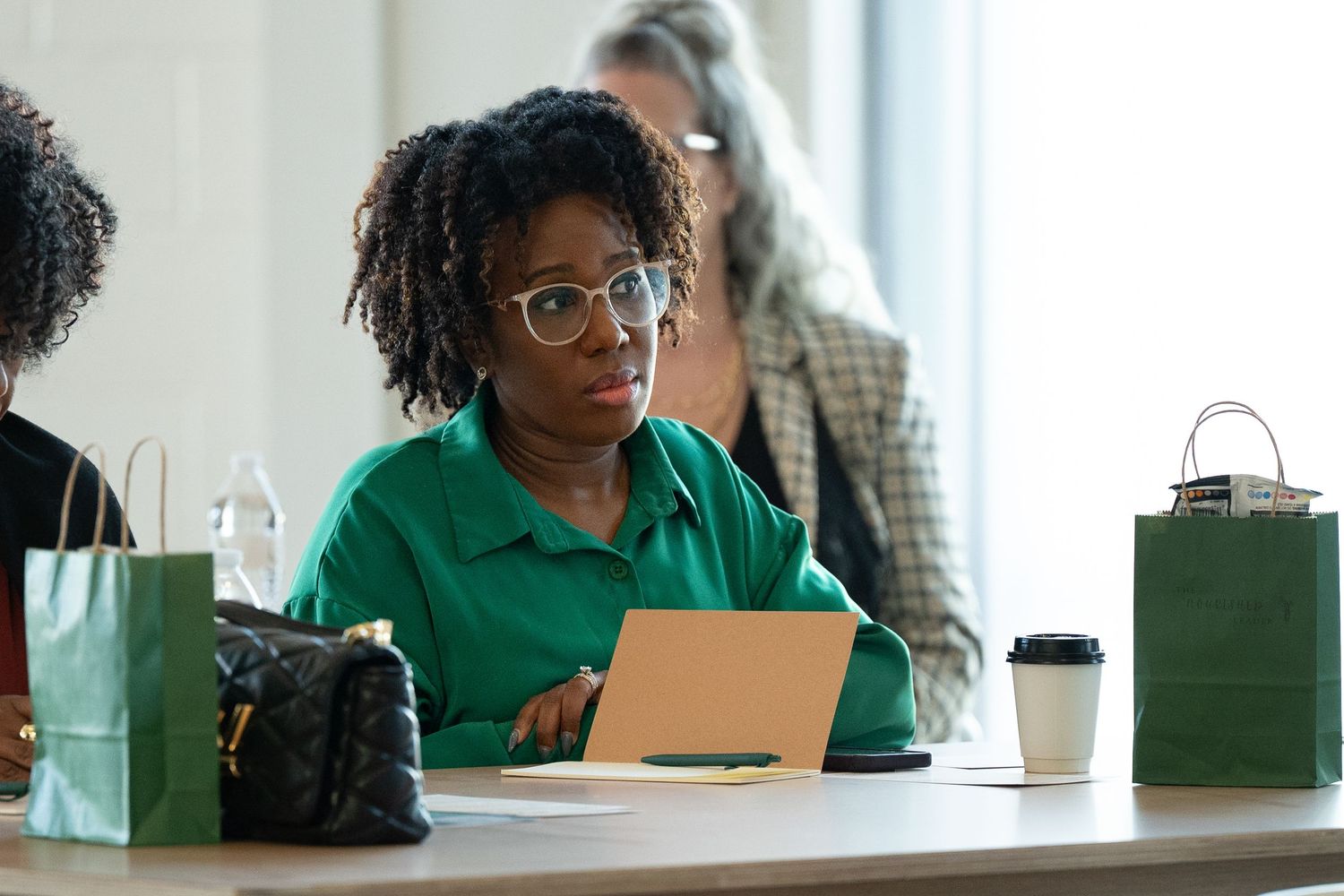 Woman in green shirt wearing glasses writing notes at a table with green gift bags.