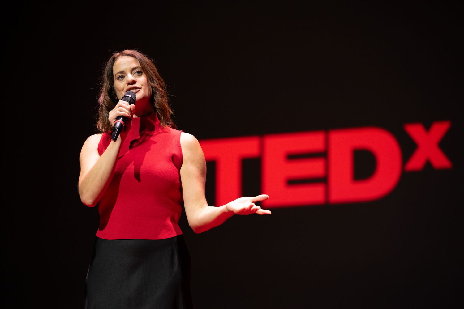 Woman in red sleeveless top holding microphone while presenting at TEDx event on stage.