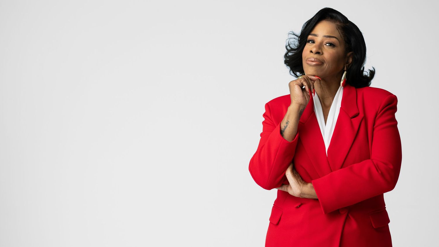 A confident woman in a red blazer poses thoughtfully against a light gray background.