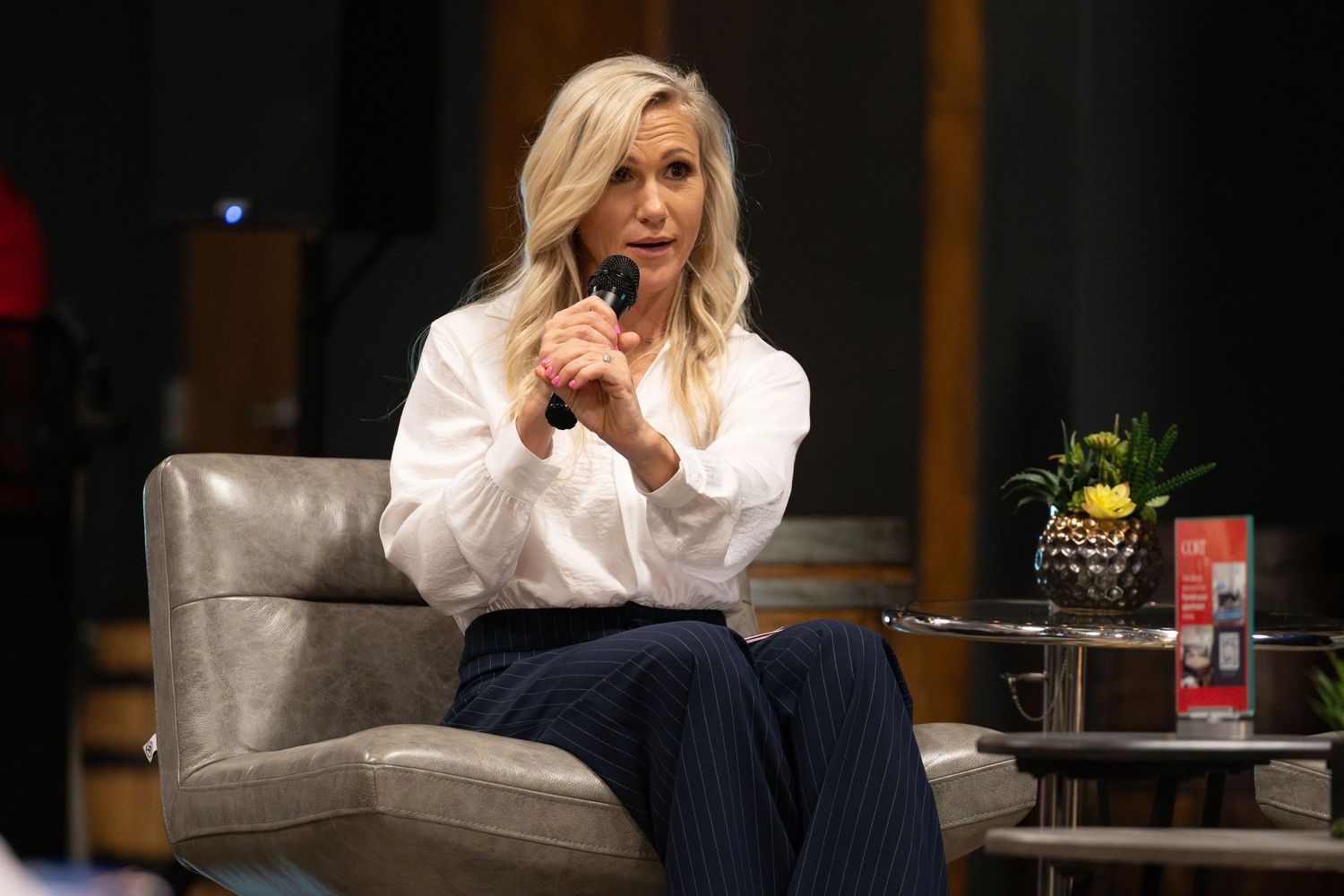 Blonde woman in white blouse and navy pinstripe pants speaks into microphone while seated in gray chair on stage.