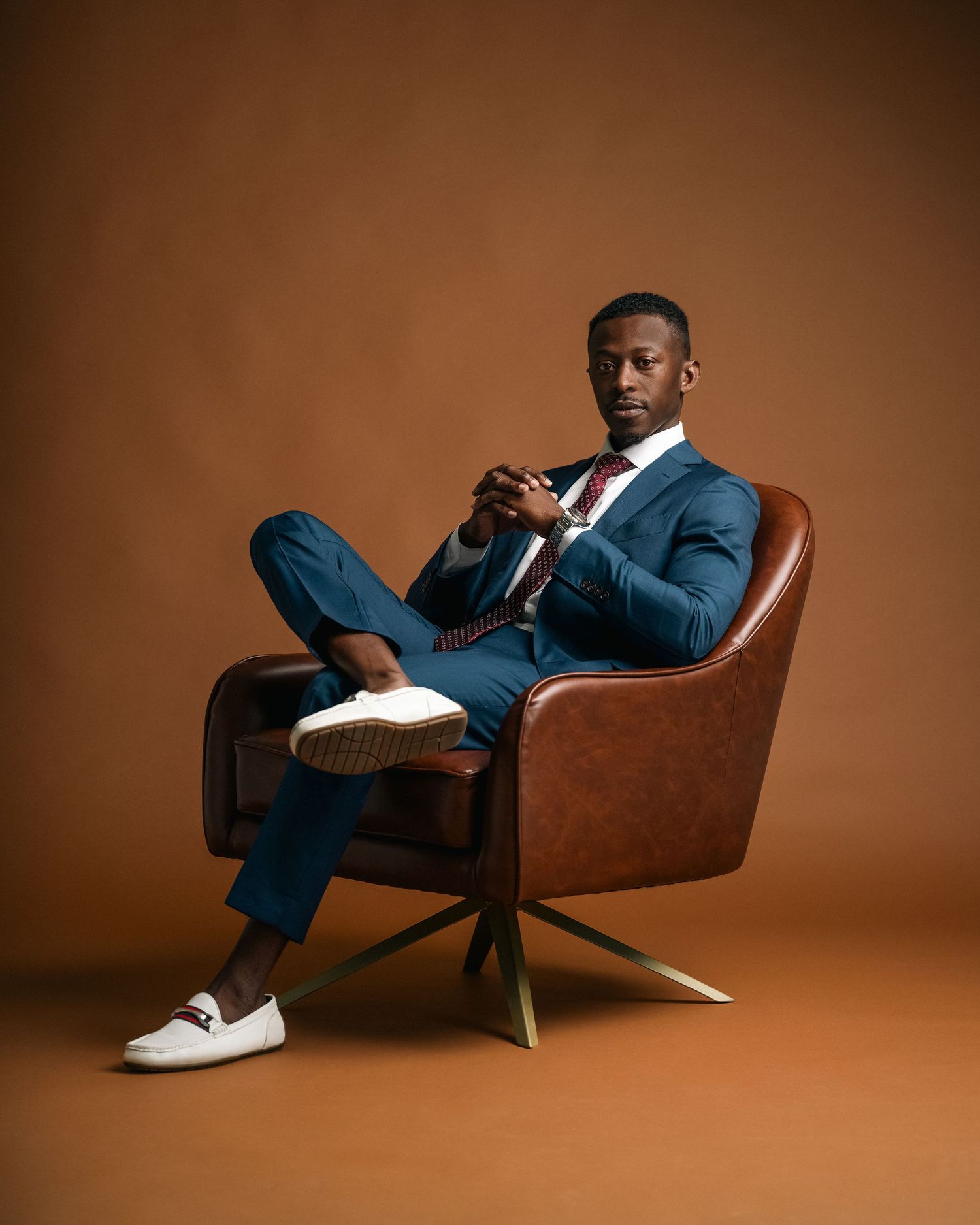 A well-dressed man in a blue suit sits confidently in a brown leather chair against a warm tan background.