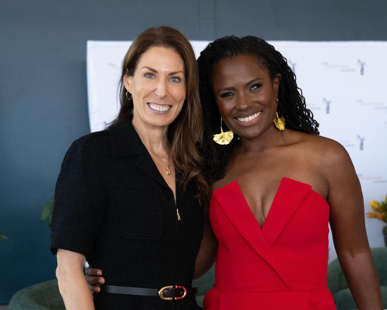 Two women posing together smiling, one in black outfit and one in red strapless dress.