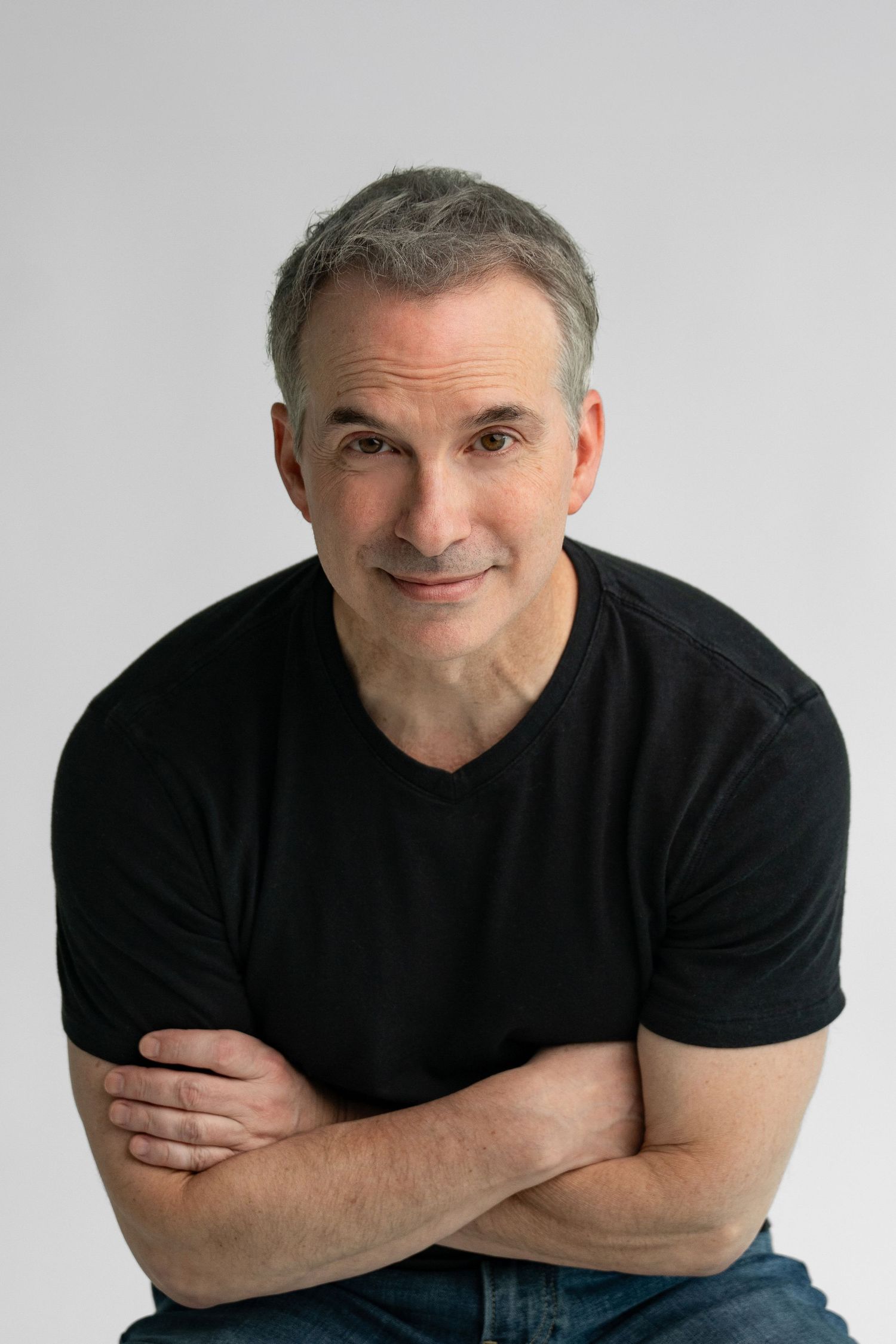 Middle-aged man with gray hair in black t-shirt sits with arms crossed, smiling confidently against white background.