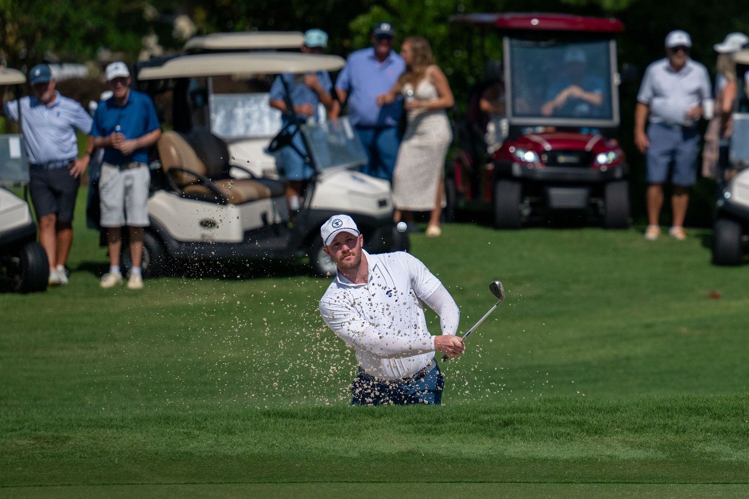 Golfer hits shot from sand bunker during tournament with spectators and golf carts in background.