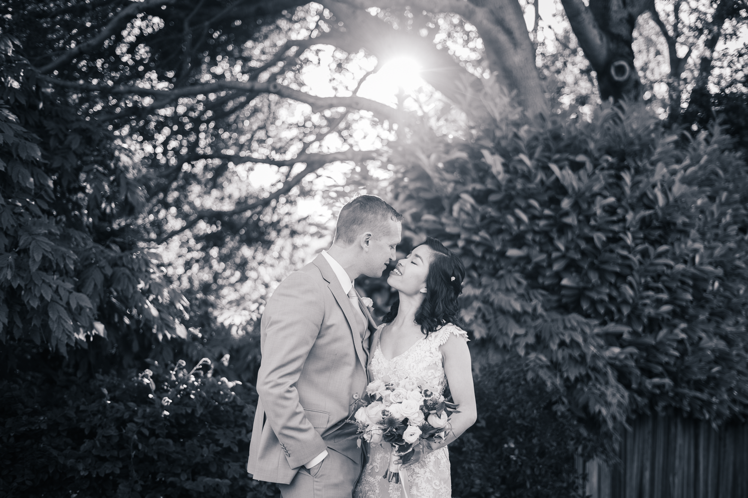 A romantic black and white wedding photo of a couple embracing in a garden with sunlight filtering through trees.