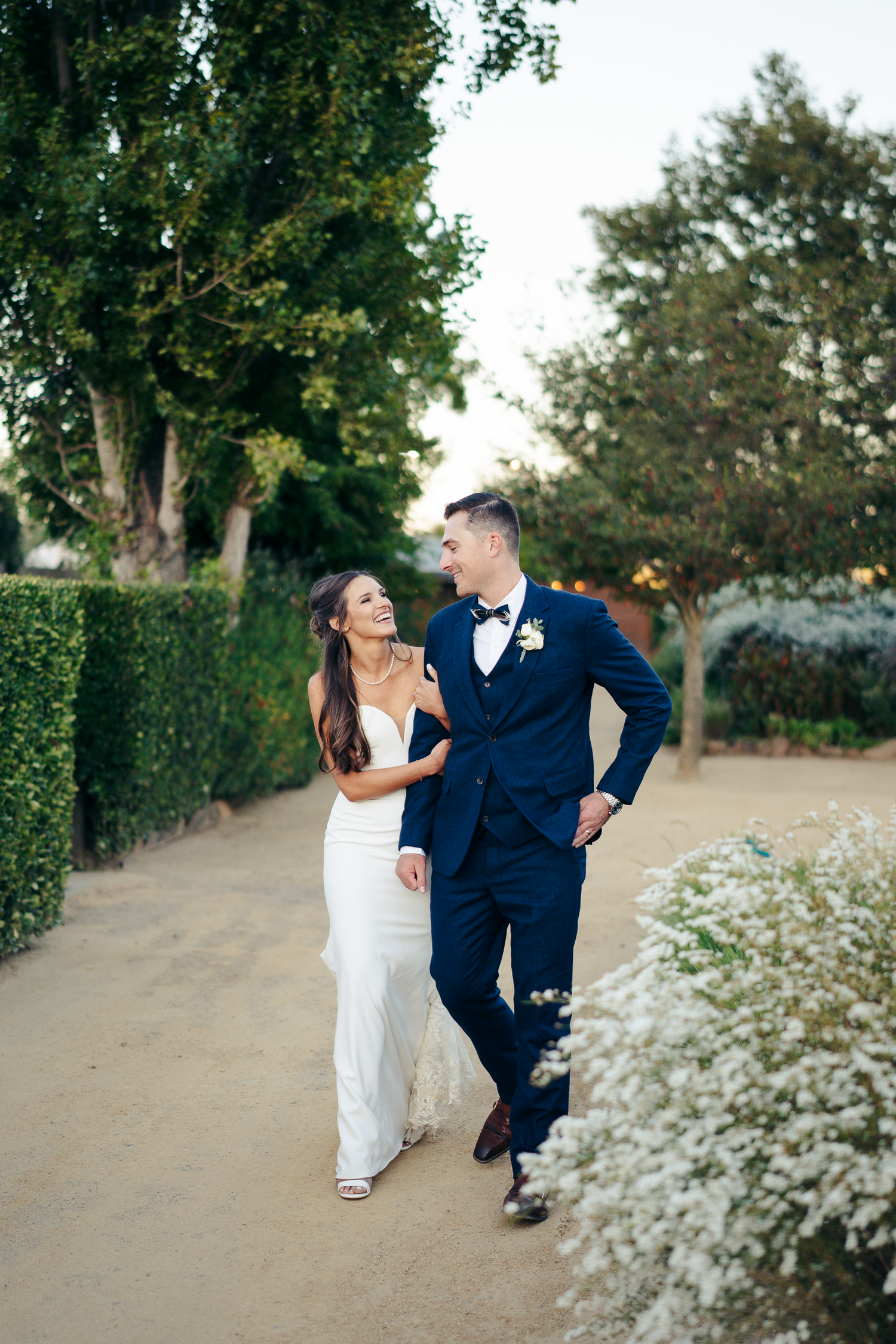 A couple in wedding attire walking together along a garden path lined with white flowering bushes at Cornerstone Sonoma.