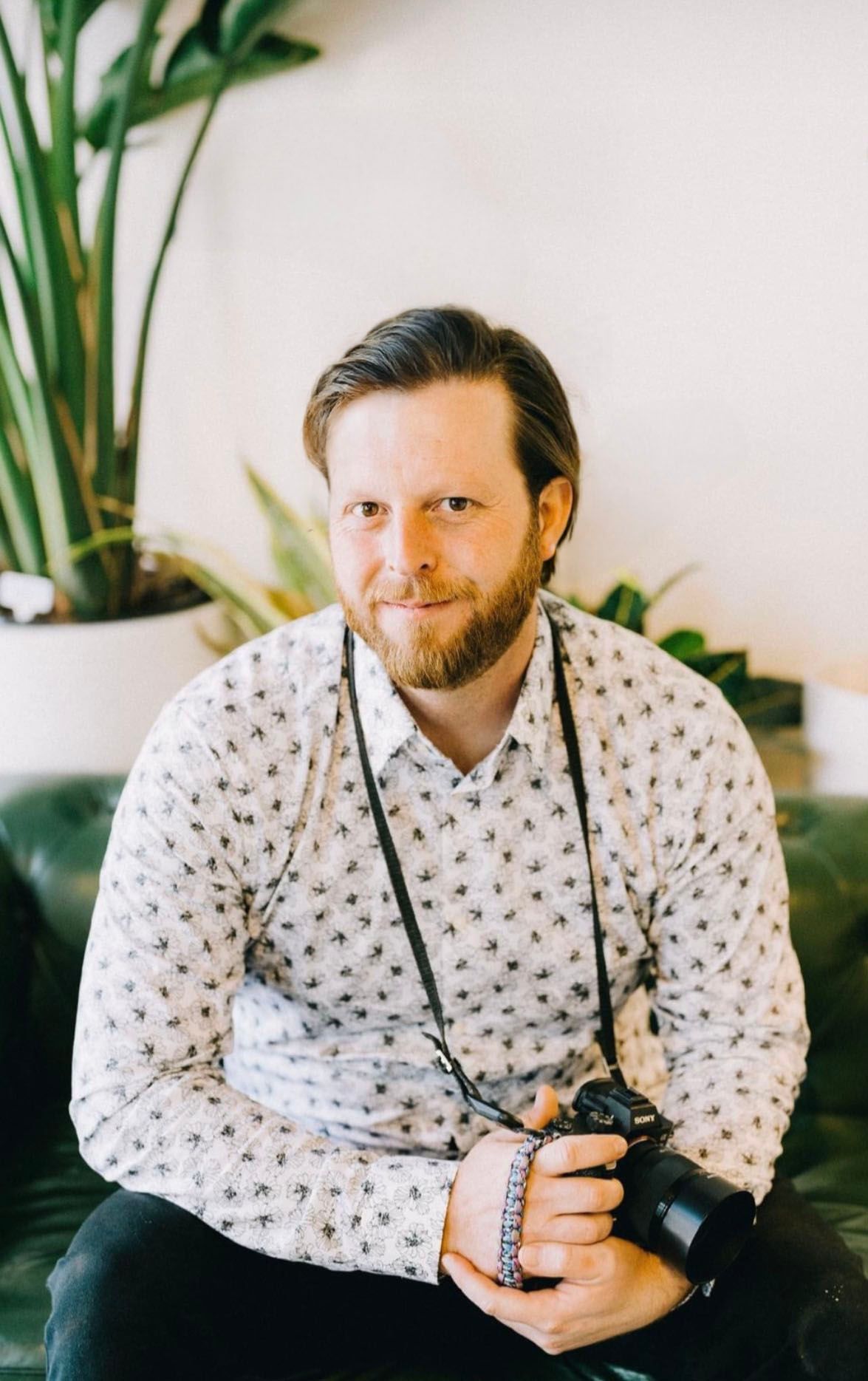 Wedding photographer, Samuel Henderson, in a patterned shirt smiling while holding professional camera equipment.
