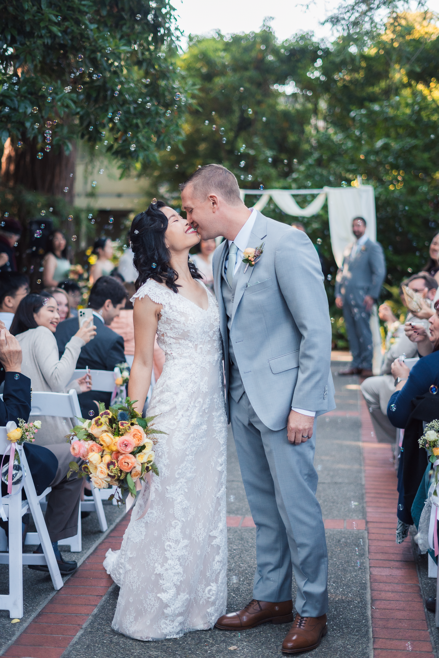 A couple shares a romantic moment during an outdoor garden wedding ceremony with guests seated on white chairs.