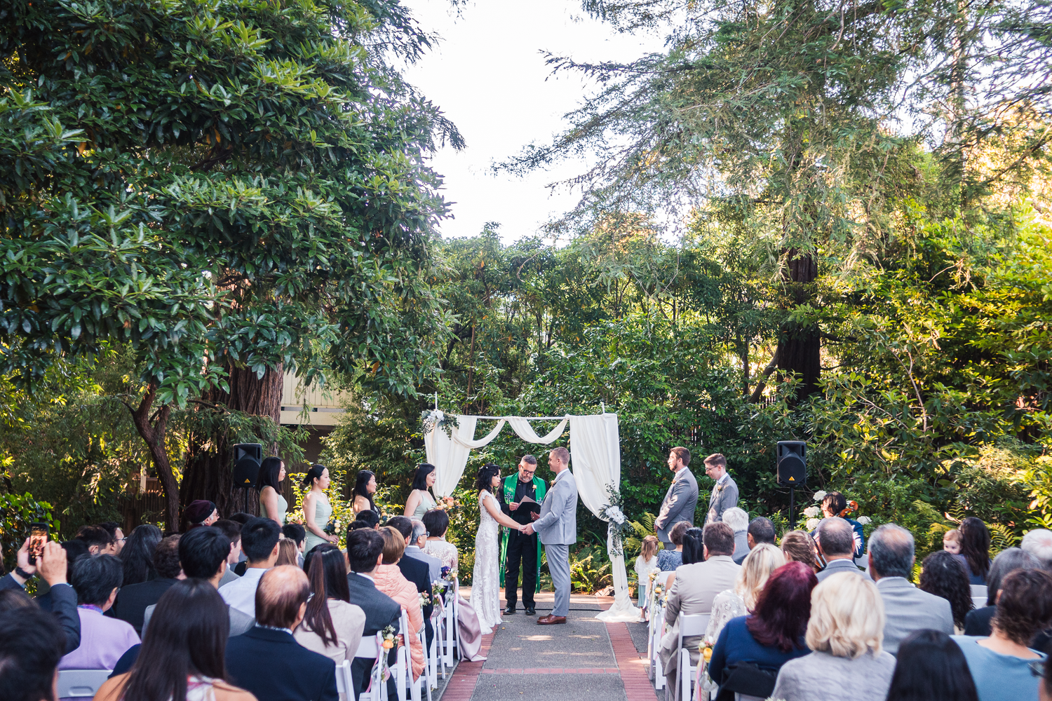Outdoor wedding ceremony in a garden setting with guests seated in white chairs beneath tall trees at sunset.