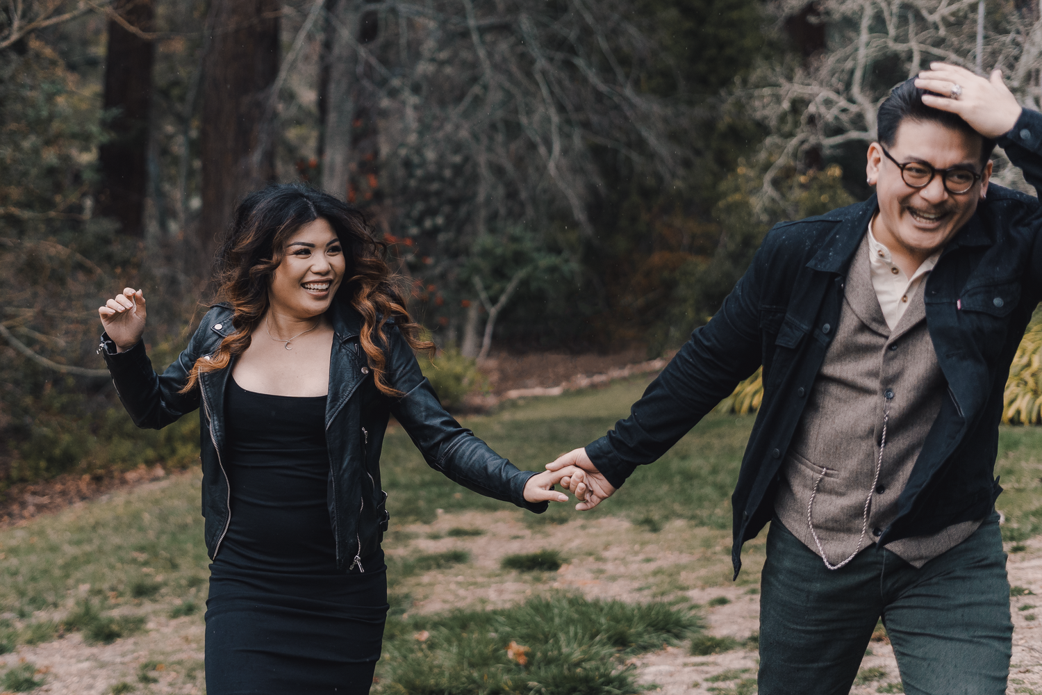 Two people holding hands and laughing while running together outdoors in a wooded area.