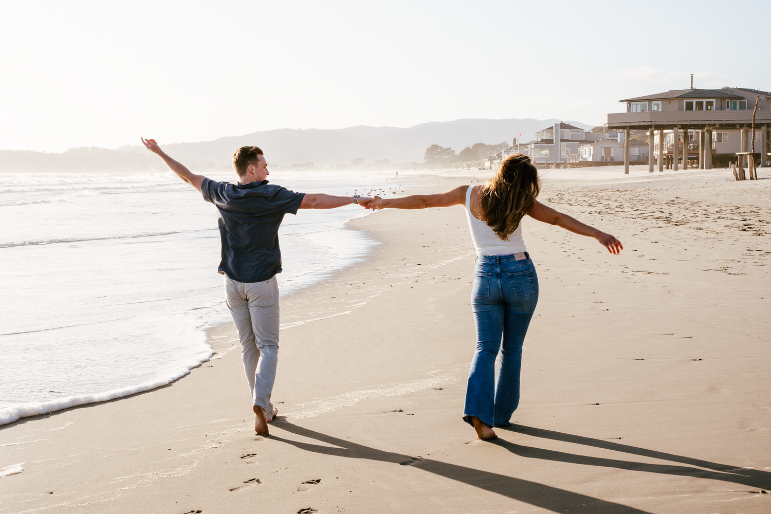 Playful couple walking with outstretched arms along a sunny beach shoreline at Stinson Beach in Marin County.