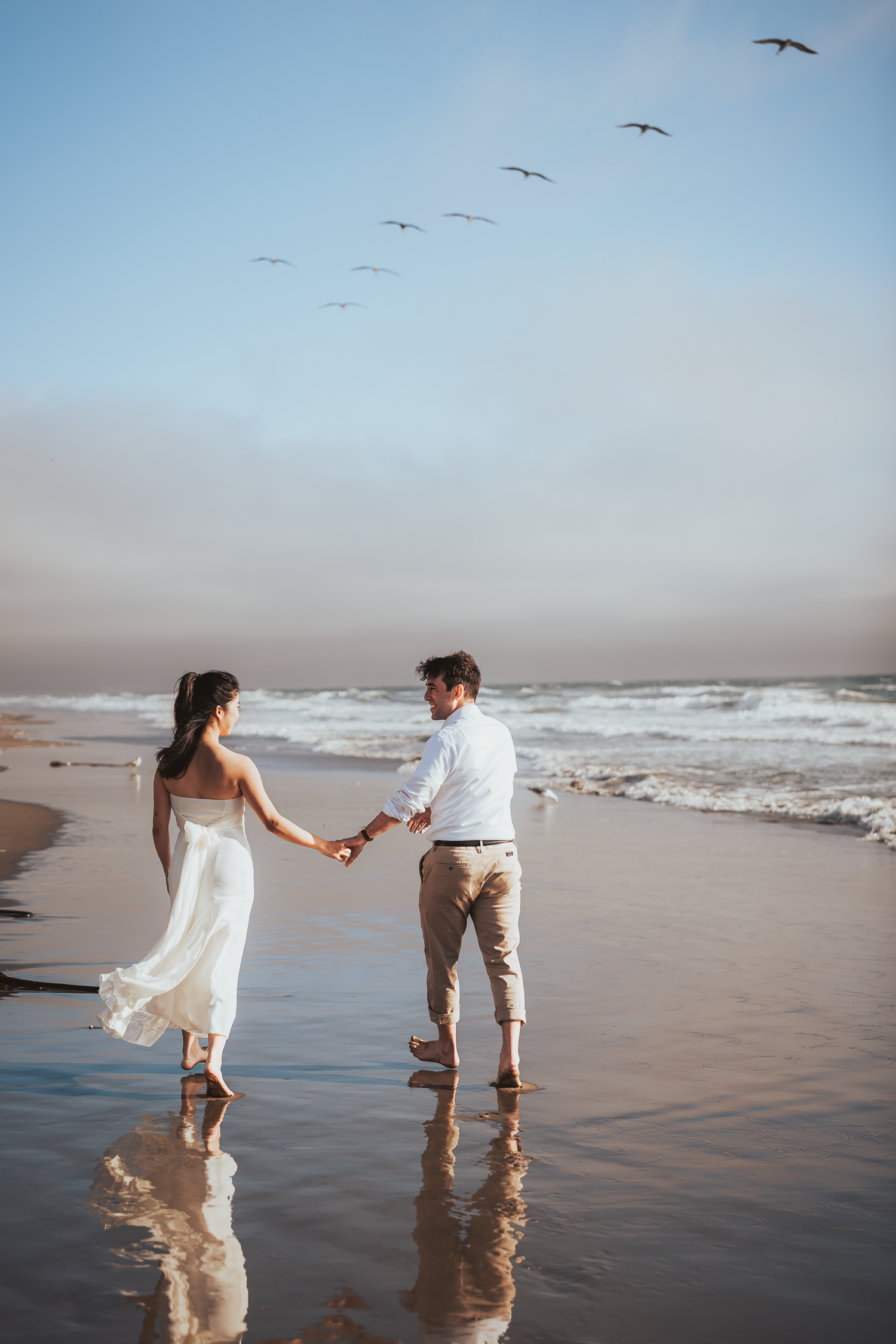 Two people hold hands and play along the shoreline as seagulls fly overhead at Ocean Beach in San Francisco.