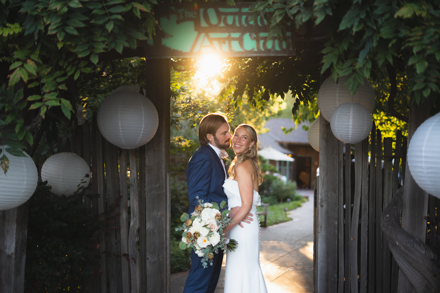Newlyweds share romantic kisses at sunset under glowing paper lanterns in an outdoor garden setting.