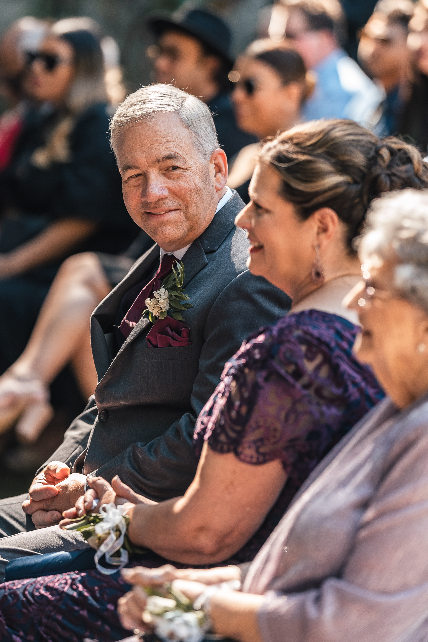 Wedding guests in formal attire smile and watch the ceremony from their seats.